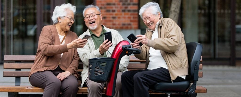 Drie lachende senioren zitten buiten op een bankje te lachen en houden een smartphone vast. Eén persoon zit op een scootmobiel. Ze lijken samen te genieten van online vermaak op hun telefoons.