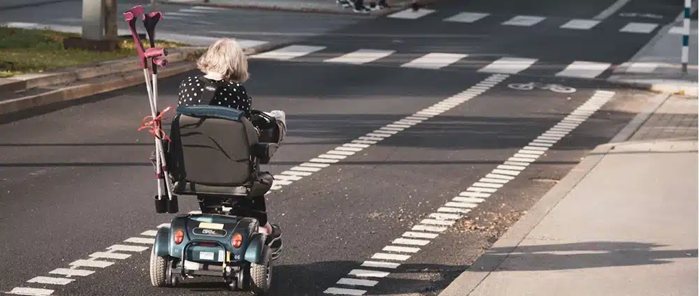 Een oudere persoon met grijs haar rijdt in een scootmobiel op een straat in de stad, met krukken achterop - een veelvoorkomend tafereel voor mensen die overwegen scootmobiel te kopen of kijken naar mogelijkheden om scootmobiel te financieren. De fietser rijdt in de richting van een oversteekplaats naast het fietspad.